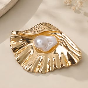 May include: A gold-toned metal brooch shaped like an open seashell with a textured, rippled surface. A single, large, irregular white baroque pearl is set in the center of the shell, creating a focal point against the polished metallic finish.