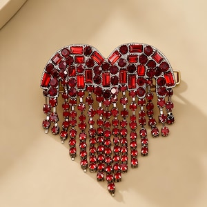 May include: A heart-shaped hair clip featuring a dark metal base encrusted with numerous red crystals in various shapes and sizes. Multiple strands of red crystal-adorned chains hang from the bottom edge, creating a cascading fringe effect.