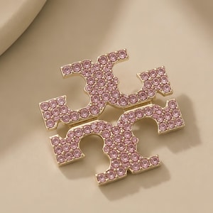 May include: A gold-toned metal brooch featuring the Tory Burch double T logo, embellished with numerous small, round, light pink crystals set across the entire surface of the emblem, creating a sparkling and reflective appearance on the metal.