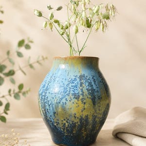 Peut inclure: Un vase en céramique au corps arrondi et à large ouverture, doté d'un émail réactif dans des tons de bleu, vert et jaune avec une finition texturée et mouchetée. Le vase contient un bouquet de tiges fleuries délicates vert clair.