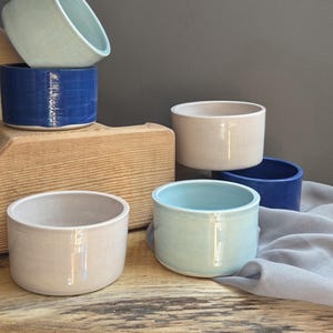 May include: A collection of six cylindrical ceramic bowls in various colors including light blue, deep blue, and off-white. The bowls have smooth, glossy surfaces and straight sides, arranged in a cluster with some stacked and others placed individually.