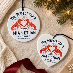 Könnte beinhalten: Zwei weiße Keramikornamente, eines rund und eines herzförmig, mit einer roten Hummerillustration, die ein Herz bildet, einem goldenen Diamantring und blauem Text THE PERFECT CATCH, MIA & ETHAN, ENGAGED, AUGUST 22, 2026, mit blauen Sternen.