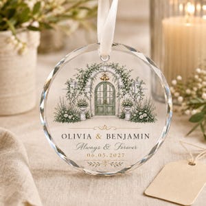 Puede incluir: Un adorno circular de cristal transparente con una cinta blanca, que presenta una ilustraci&oacute;n de una puerta arqueada verde rodeada de enredaderas florales y campanas doradas. El texto dice OLIVIA & BENJAMIN, Always & Forever, 06.05.2027.