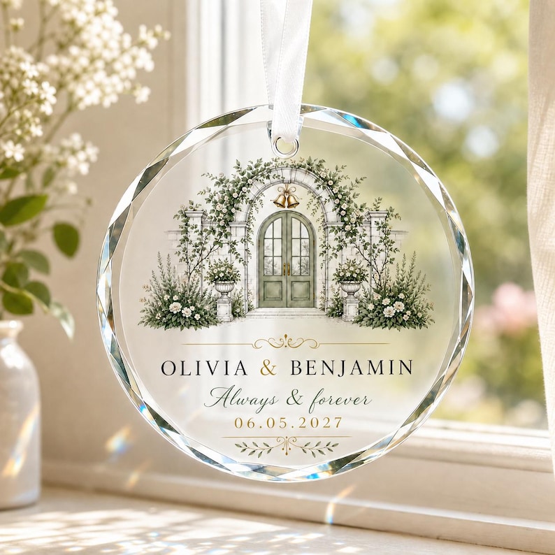 Puede incluir: Un adorno de cristal circular y facetado que cuelga de una cinta blanca. El centro presenta una ilustraci&oacute;n impresa de un arco de piedra con puertas verdes, campanas doradas y arreglos florales. El texto dice: OLIVIA & BENJAMIN, Always & forever, 06.05.2027.