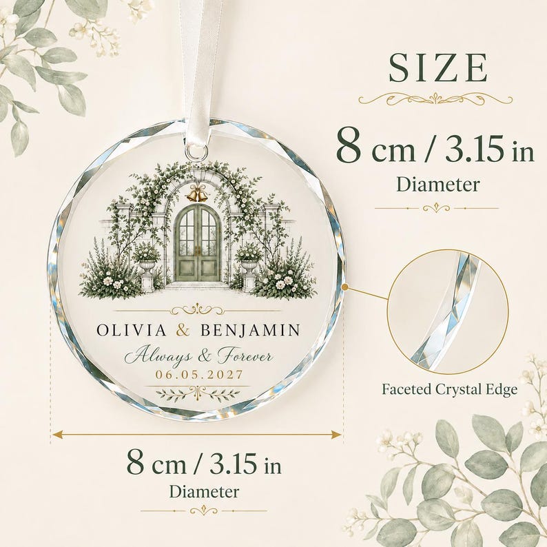Puede incluir: Un adorno redondo de cristal con borde facetado, que presenta una ilustraci&oacute;n impresa de un arco floral y puertas dobles. Incluye el texto OLIVIA & BENJAMIN, Always & Forever y 06.05.2027. El adorno tiene un di&aacute;metro de 8 cm y una cinta blanca.
