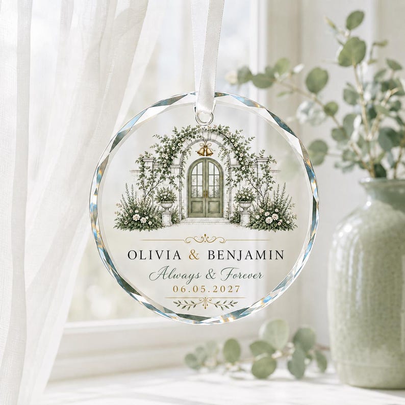 Puede incluir: Un adorno circular de cristal transparente con bordes facetados, que presenta una ilustraci&oacute;n de un arco de jard&iacute;n con puertas verdes y flores. El texto dice OLIVIA & BENJAMIN, Always & Forever y 06.05.2027, suspendido por una cinta blanca.