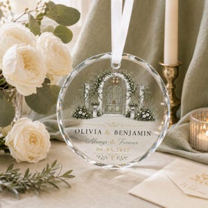 Puede incluir: Un adorno circular de cristal transparente con una cinta blanca, que presenta una ilustraci&oacute;n de un arco de piedra con flores. El texto dice OLIVIA & BENJAMIN, Always & Forever, y 06.05.2027 en letras negras y doradas.