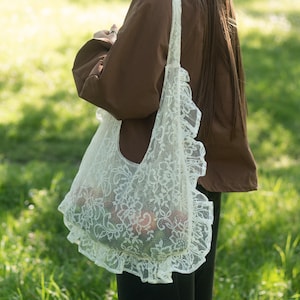 Puede incluir: Un bolso de mano de encaje blanco con un patrón floral y bordes con volantes en el lateral. El bolso tiene una sola correa para el hombro y una estructura suave y semitransparente, mostrando un delicado diseño calado en toda la tela.