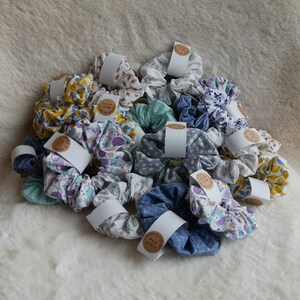 Könnte beinhalten: Eine Sammlung von Haargummis aus Stoff in verschiedenen Farben und Mustern, darunter Blumen-, geometrische und Sternenprints. Jedes Haargummi ist mit einem weißen Papierband befestigt, das ein rundes Etikett mit Handmade with love trägt.