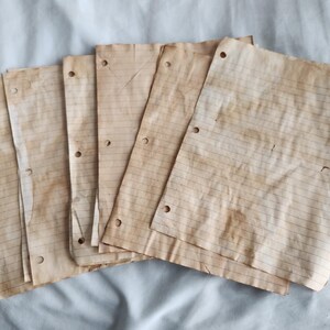 May include: A stack of aged, lined paper sheets with three-hole punches. The paper has a warm, sepia tone with visible staining and wear, suggesting a vintage or antique aesthetic. The horizontal lines are evenly spaced.