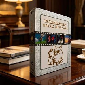 May include: A white and silver box set titled The Collected Works of Hayao Miyazaki. The front features a film strip graphic with colorful scenes and a line drawing of a pig wearing sunglasses at a desk. The textured cover has a metallic finish.