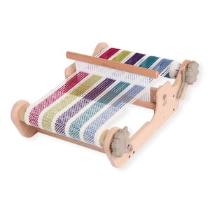 Op de afbeelding: Een kleine houten weefgetouw met kleurrijk garen in tinten rood, groen, paars en blauw. Het weefgetouw heeft een houten frame en verstelbare knoppen. De geweven stof vertoont een gestreept patroon.