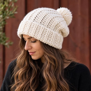 May include: A cream-colored knit beanie featuring a thick ribbed texture, a wide folded brim, and a large matching yarn pom-pom on the top. The hat is constructed with a chunky yarn pattern that creates a distinct vertical ribbed appearance.