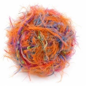 Può includere: Un gomitolo di filato testurizzato con un mix vivace di fibre arancioni, rosa, blu e verdi. Il filato ha un aspetto irregolare e soffice con spessori variabili e estremità libere che creano un effetto morbido, voluminoso e multicolore.