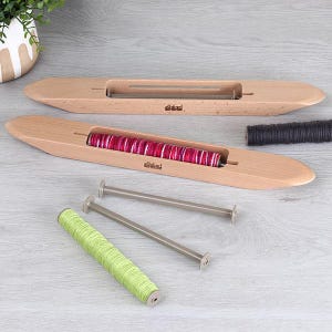 Op de afbeelding: Twee houten weefshuttles met een lichte houtafwerking. Eén shuttle bevat een spoel roze en wit garen, de andere is leeg. Extra spoelen groen en zwart garen zijn ook aanwezig, samen met twee metalen spoelen. De shuttles zijn voorzien van de merknaam.