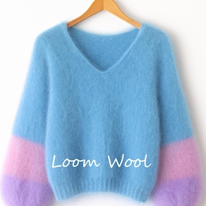 Può includere: Un maglione blu chiaro con scollo a V e maniche lunghe. Le maniche sono a blocchi di colore con sezioni rosa e viola. Il testo "Loom Wool" è scritto in bianco sul davanti. Il maglione è appeso a una gruccia di legno.