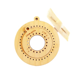 Op de afbeelding: Een ronde breimachine van hout met een gat in het midden en een verscheidenheid aan verschillende gaten rond de buitenrand. De breimachine is gelabeld "LOOM-WOOL" en heeft de nummers 1, 1.4, 2, 2.19, 3 en 6 gedrukt rond de buitenrand.