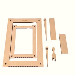 Puede incluir: Un kit de telar de madera con dos marcos rectangulares, dos agujas, una herramienta en forma de tenedor y dos piezas rectangulares. Los marcos tienen bordes dentados y el texto "LOOM-WOOL". Las herramientas son de madera clara.