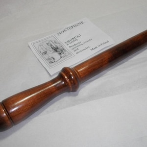 Peut inclure: Un pédalier en bois pour roue à filer, également appelé "nostep pinne", fabriqué par Kromski & Sons en Pologne. Le pédalier est brun et a une extrémité arrondie.