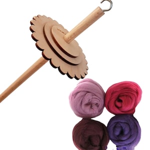 Op de afbeelding: Een houten spinnewiel met een bloemvormige basis en een haak aan de bovenkant. Vier bollen wol in tinten roze, paars en bruin bevinden zich op de voorgrond.