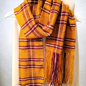 May include: Mustard yellow scarf with a plaid pattern in burgundy and white. The scarf has fringing at both ends and is displayed on a white hanger. The scarf is made of woven fabric.