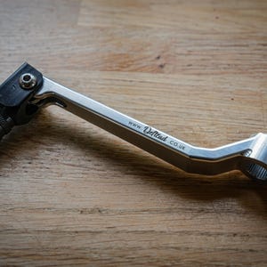 Pode incluir: Uma alavanca de mudanças de motocicleta em metal prateado com um apoio para os pés preto serrilhado, uma articulação com um parafuso prateado e uma braçadeira de montagem com um parafuso. O braço da alavanca tem o texto www.deftcad.co.uk gravado.