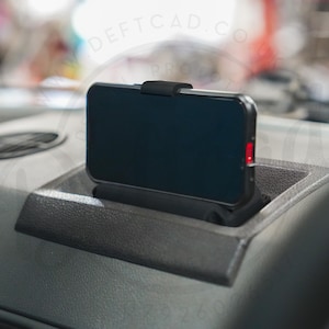Könnte beinhalten: Ein schwarzes Smartphone in einer passgenauen schwarzen Kunststoffhalterung, die in einem vertieften Armaturenbrettfach befestigt ist. Das Gerät ist horizontal ausgerichtet, hat einen dunklen Bildschirm und ein kleines rotes Licht an der Seite.