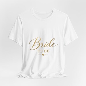Könnte beinhalten: Ein weißes Kurzarm-T-Shirt mit dem goldfarbenen Schriftzug "Bride TO BE" mittig auf der Brust. Unter dem Text befindet sich ein kleines goldenes Herz, das von zwei horizontalen Linien flankiert wird. Das Shirt hat einen Rundhalsausschnitt.