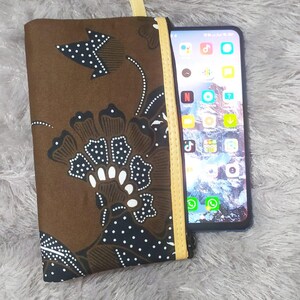 Op de afbeelding: Een rechthoekig stoffen tasje met een bruin en zwart bloemrijk batikpatroon, witte stippendetails en een gele ritssluiting. Een smartphone met een scherm dat diverse kleurrijke app-iconen weergeeft, is naast het tasje geplaatst.
