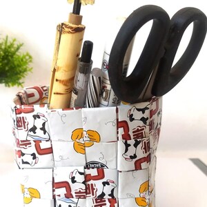 May include: A square pencil holder crafted from woven white paper strips featuring a repeating pattern of black and white cows, yellow circular shapes, and red Korean text. The container holds a pair of black scissors and several writing pens.