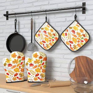 May include: A kitchen set featuring two square potholders and two oven mitts with a white background and a colorful autumn leaf pattern in shades of orange, yellow, red, and green. A black frying pan and a black spatula hang on a wall-mounted rack.