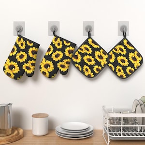 May include: Two oven mitts and two square pot holders featuring a repeating pattern of yellow sunflowers on a black background. Each item has a black fabric trim and a small loop for hanging. The set is displayed hanging on four wall hooks.