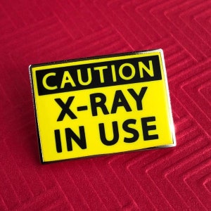 May include: Yellow and black enamel pin with the text "CAUTION X-RAY IN USE" on a red background.