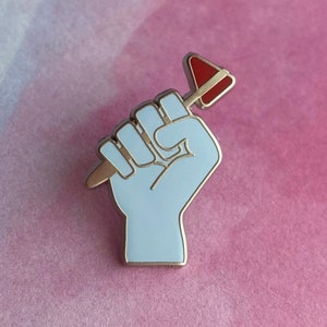 Puede incluir: Un pin de esmalte plateado con un puño en alto sosteniendo un martillo rojo. El puño es azul claro y el martillo es rojo.
