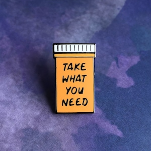 Puede incluir: Pin de esmalte de forma de píldora naranja con contorno negro. El pin dice "Toma lo que necesitas".