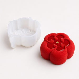 Könnte beinhalten: Eine durchscheinende, blumenförmige Silikonform steht neben einem fertigen, leuchtend roten Dessert mit samtiger Textur in Form einer fünfblättrigen Blüte. Das Dessert hat eine glatte, glänzende rote Mitte mit einem kleinen Blattgoldstück.