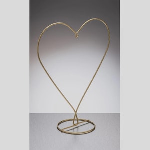 May include: A gold-toned metal display stand shaped like a heart. The structure consists of a thin wire frame forming a heart outline, featuring a small hook at the top center for hanging items, supported by a circular base made of the same metal.