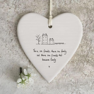 Op de afbeelding: Een wit, hartvormig keramieken hangbordje met een zwarte lijntekening van twee huizen en een boom. De tekst luidt: There are friends, there are family and there are friends that become family. Onderaan bevindt zich een kleine witte bloem.