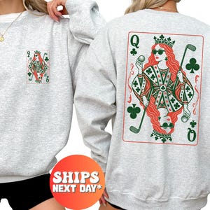May include: A light gray crewneck sweatshirt featuring a graphic of a playing card queen wearing sunglasses and holding a golf club and ball. The design is printed in red and green on the back and in a smaller size on the left chest.