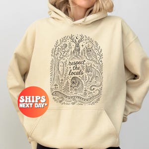 May include: A beige hooded sweatshirt featuring a black ink-style illustration of various forest creatures including a rabbit, owl, and lizard, surrounded by trees and foliage, with the text "respect the locals" printed in the center.