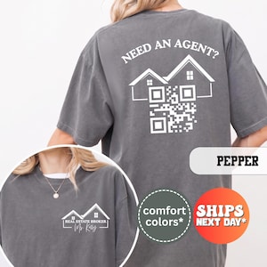May include: A charcoal gray t-shirt featuring a white graphic on the back with the text "NEED AN AGENT?" above a house outline and a QR code. The front chest area displays a smaller white house graphic with the text "REAL ESTATE BROKER Ms. Kelly".