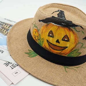 May include: A tan woven fedora hat featuring a hand-painted orange jack-o'-lantern wearing a black witch hat, green leaves, and a small black bat. The hat has a wide brim and a solid black ribbon band encircling the base of the crown.