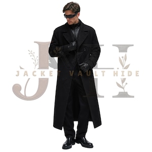May include: A long black wool-blend overcoat featuring a wide notched lapel, double-breasted button closure, and structured shoulders. The coat is worn over a black zip-up jacket, black trousers, black leather gloves, and black shoes.