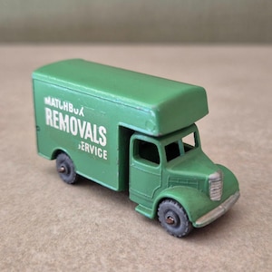 Op de afbeelding: Een kleine groene gegoten speelgoedvrachtwagen met een doosvormige laadruimte. Op de zijkant van de vrachtwagen staat de witte tekst MATCHBOX REMOVALS SERVICE. Het voertuig heeft een zilverkleurige grille en bumper met kleine grijze wielen.