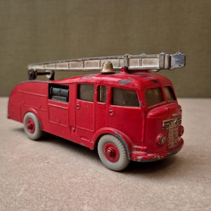 Op de afbeelding: Een vintage rode speelgoedbrandweerwagen met een zilverkleurige ladder op het dak, een kleine bel en lichtgrijze wielen. De metalen carrosserie vertoont gebruikssporen met afgebladderde verf en de grille aan de voorzijde heeft een metallic afwerking.