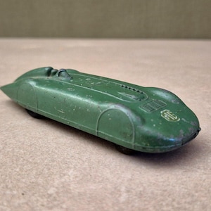 May include: A vintage green die-cast toy racing car with a streamlined, elongated body. The vehicle features a small open cockpit, a textured bonnet vent, and a white circular logo on the front nose. The paint shows signs of wear and age.