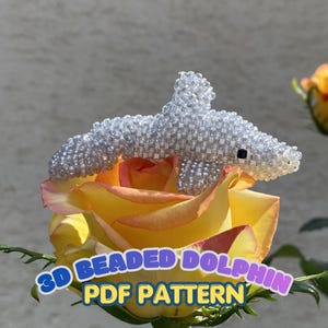 May include: A three-dimensional dolphin figure crafted from small, clear, iridescent beads, featuring a single black bead for the eye. The text 3D BEADED DOLPHIN PDF PATTERN is displayed in blue and yellow letters at the bottom of the image.
