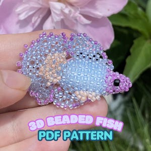 May include: A 3D beaded fish ornament crafted from small light blue, lavender, and peach-colored glass beads. The fish features a rounded body, a prominent tail fin, and a single black bead for an eye. The text 3D BEADED FISH PDF PATTERN is visible.