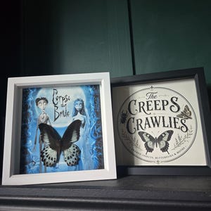 May include: Two square shadow box frames. The white frame contains a Corpse Bride movie print with a mounted black and white butterfly. The black frame features a circular logo reading The Creeps Crawlies Framed Insects, Butterflies & Moths.