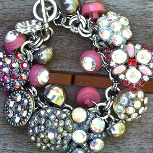 May include: A silver charm bracelet with pink, red, and clear rhinestone buttons. The bracelet has a silver clasp and is decorated with various charms, including hearts, flowers, and stars.